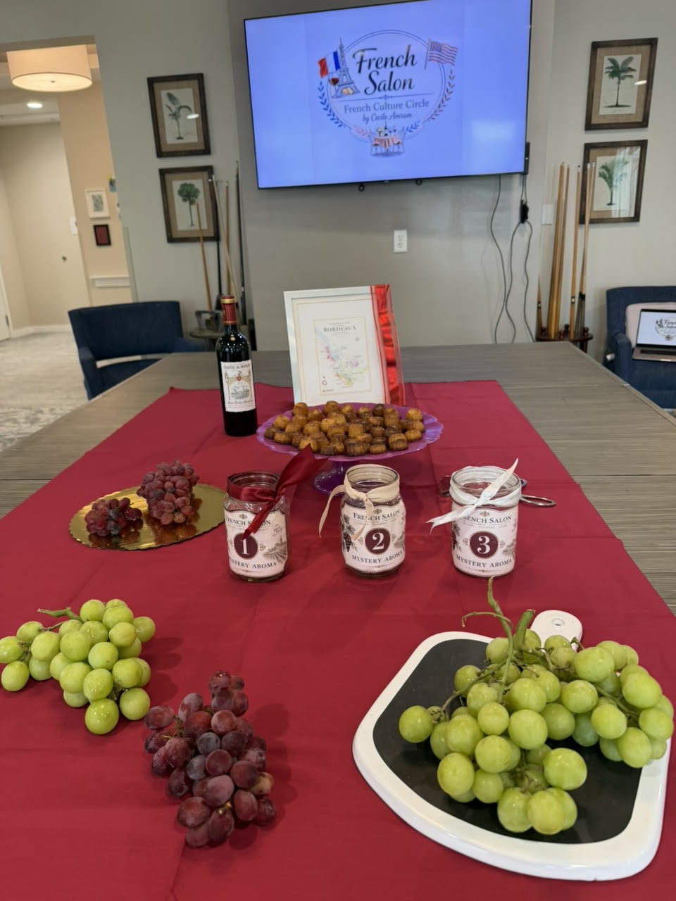 A red tablecloth set with grapes, canelés, a bottle of wine, and three French Salon mystery aroma jars.