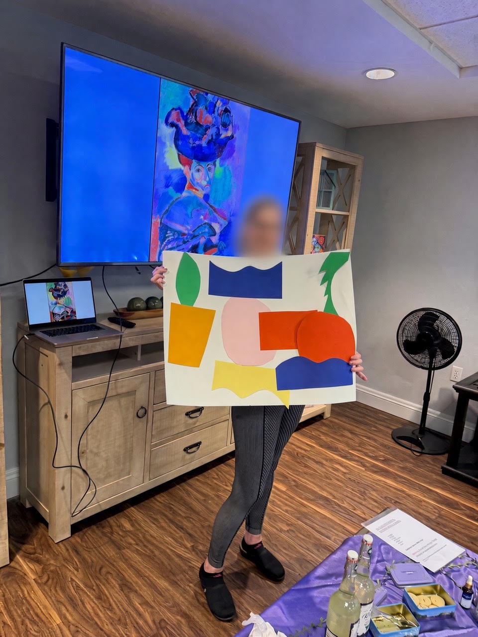 A facilitator holding a colorful paper cut-out in front of a screen showing a Matisse portrait.